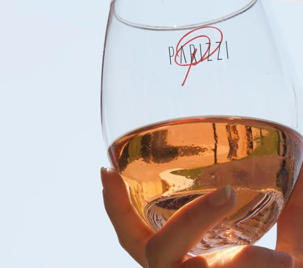 Un calice di vino rosato alzato controluce contro il cielo, con il logo Parizzi inciso sul vetro