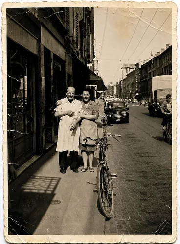 Foto d'epoca in strada, davanti al negozio, con biciclette e automobili dell'epoca