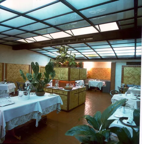 Sala degli anni Ottanta con la copertura a vetri e le piante fra i tavoli